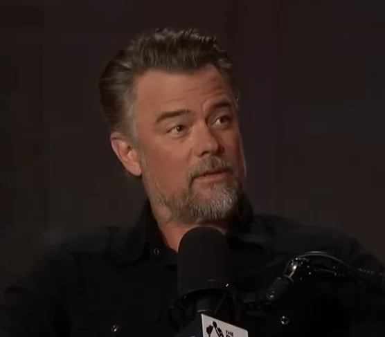 Man with grey hair and facial hair speaking into a microphone against a dark background