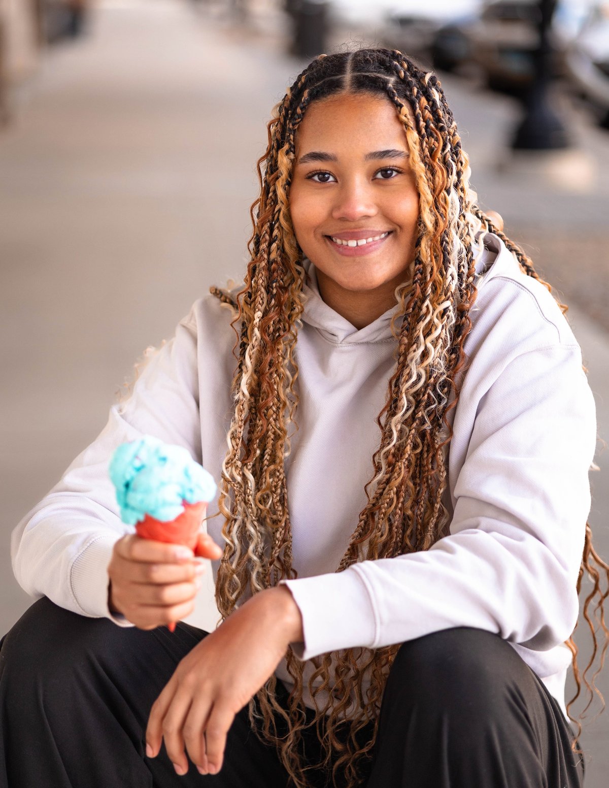 Young woman with braided hair smiling at camera, wearing white hoodie and holding colorful popsicle