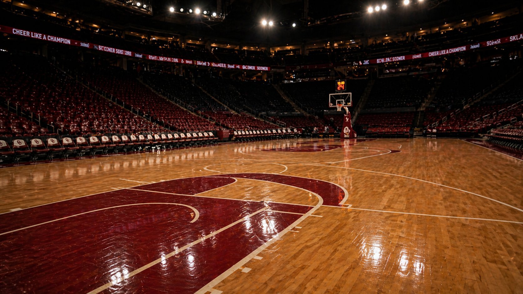 Empty NBA basketball arena with polished wooden court, red and gold center logo, basket at far end, and packed seating sections under bright arena lights